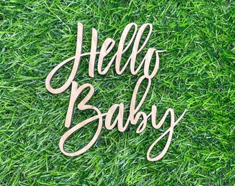 May include: Wooden sign with the words "Hello Baby" in a cursive font. The sign is on a green grass background.