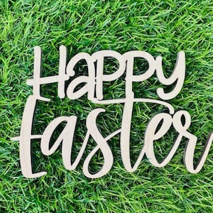 May include: A wooden sign with the words "Happy Easter" in a cursive font. The sign is light-colored and sits on a bed of green grass. The letters are connected, creating a decorative Easter-themed item.