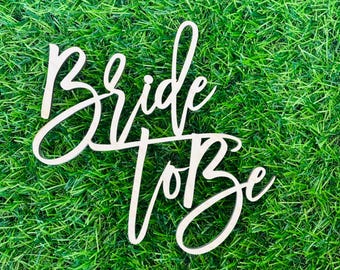 May include: White wooden sign with the text "Bride to Be" in cursive script. The sign is on a green grassy background.