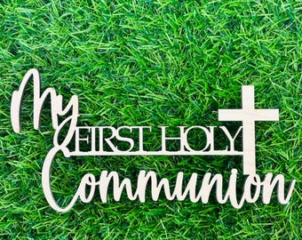 May include: Wooden sign with the text "My First Holy Communion" in white lettering on a natural wood background. The sign is on a green grass background.