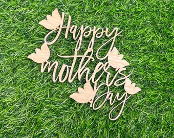 Könnte beinhalten: Holzschild mit dem Schriftzug "Happy Mother's Day" in geschwungener Schrift, verziert mit hölzernen Blumenmotiven. Präsentiert auf einem Hintergrund aus grünem Gras.