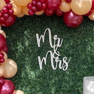 May include: White wooden "Mr & Mrs" sign with cursive lettering against a backdrop of green artificial grass and a balloon arch with burgundy and beige balloons.