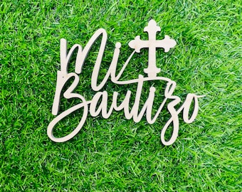 Könnte beinhalten: Ein Holzschild mit dem Text "Mi Bautizo" und einem Kreuz in kursiver Schrift. Das Schild befindet sich auf einem grünen Gras Hintergrund.