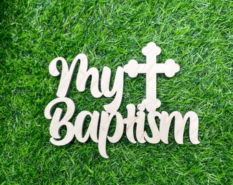 May include: A wooden sign with the words "My Baptism" in a cursive font, with a cross above the word "Baptism". The sign is made of light-colored wood and is set against a background of green grass.
