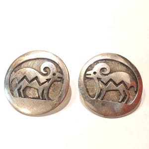 May include: A pair of circular silver earrings featuring a cutout design of a bighorn sheep. The metal has a polished finish on the outer edges and a textured, darkened background within the recessed areas of the animal motif.