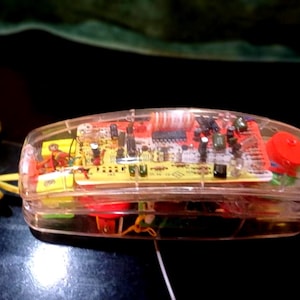 Puede incluir: Un auricular de teléfono transparente que revela sus componentes electrónicos internos. El auricular tiene un botón rojo y un cable en espiral amarillo. La placa de circuito interna es una mezcla de rojo, amarillo y verde. Probablemente un artículo de novedad o decorativo.