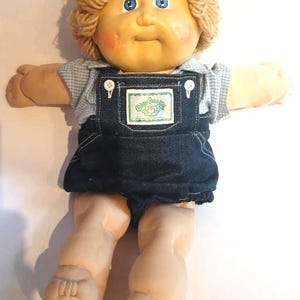 May include: A Cabbage Patch Kids doll with light skin, blue eyes, and blonde yarn hair. The doll wears a light blue and white gingham shirt under dark blue denim overalls with white buttons and a rectangular logo patch on the chest.