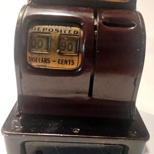 May include: A vintage coin bank, featuring a dark maroon and black color scheme. The bank has a display that reads "Deposited" and shows "00" dollars and "90" cents. The top of the bank reads "Nickels Dimes Quarters".