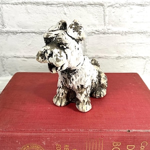 May include: A small, white and brown dog figurine with a textured appearance. The dog is sitting on a red book. The figurine has a distressed, vintage look, with visible paint wear. The background is a white brick wall.