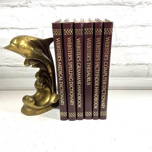 May include: A gold-colored dolphin bookend supporting a collection of Webster's reference books. The books have burgundy covers with gold lettering. Titles include Medical Dictionary, Spelling Dictionary, Grammar Handbook, Thesaurus, Factfinder Handbook, and Computer Dictionary.