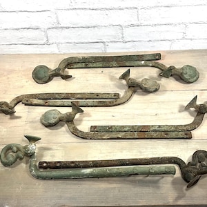 May include: A collection of antique, weathered metal hinges with decorative finials. The hinges have a curved design with a distressed, aged appearance, and are in shades of green and brown. They are arranged on a light wooden surface.