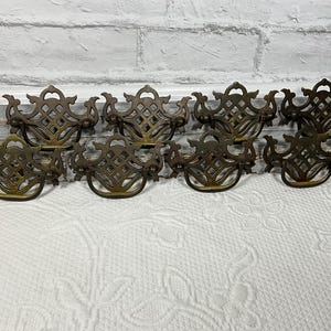 May include: A set of eight brass drawer pulls featuring an ornate, openwork lattice design with a curved, swinging handle. Each piece has a symmetrical, decorative backplate with intricate scrollwork and a polished metallic finish.