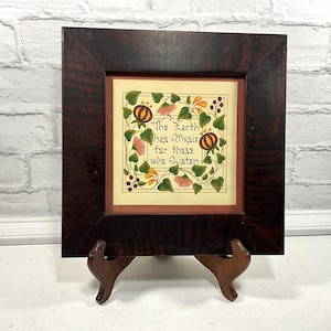 May include: A framed artwork with a dark wood frame and a cream-colored background. The artwork features the text "The Earth has music for those who listen" surrounded by floral and pumpkin illustrations. The frame is supported by a wooden easel.