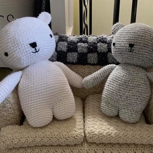 May include: Two crocheted stuffed animal bears, one white and one gray, sitting on a beige crocheted couch. The couch has a checkered pattern fabric on the back.