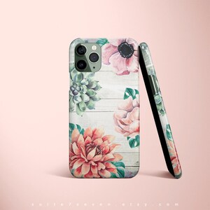 Pu&ograve; includere: Una custodia per telefono con uno sfondo in legno bianco e disegni floreali ad acquerello. I fiori sono rosa, rossi e verdi. La custodia ha un ritaglio per la fotocamera e un bordo nero.