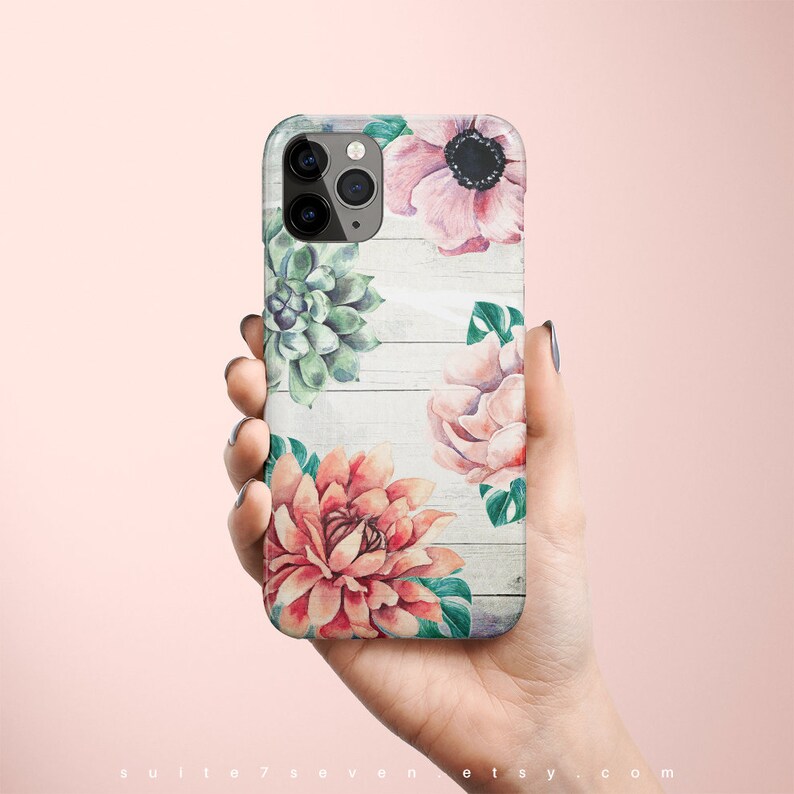 Pu&ograve; includere: Una custodia per telefono bianca con un design floreale. La custodia presenta fiori rosa, rossi e verdi su uno sfondo in legno bianco.