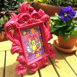 May include: A pink ornate picture frame with a purple background and a yellow cartoon character with a pink bow and stars. The frame is sitting on a wooden table next to a potted purple flower.