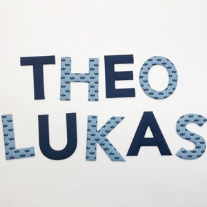 Peut inclure: Le nom "THEO LUKAS" écrit en grandes lettres bleues marine avec un motif de couronne bleu clair.
