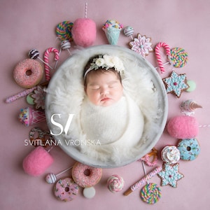 May include: A newborn baby girl wearing a white swaddle and a flower crown sleeps in a white bowl surrounded by colorful candy and donut props. The image is captioned "SVITLANA VRONSKA".