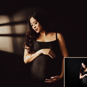 Peut inclure: Une femme enceinte portant un d&eacute;bardeur noir se tient dans une pi&egrave;ce faiblement &eacute;clair&eacute;e avec une fen&ecirc;tre derri&egrave;re elle. La lumi&egrave;re de la fen&ecirc;tre projette une ombre sur le mur derri&egrave;re elle. Elle se tient le ventre avec les deux mains.