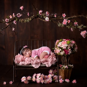 May include: A sleeping baby in a pink onesie lies on a metal bench with a pink blanket. The bench is decorated with pink flowers and a floral arrangement.