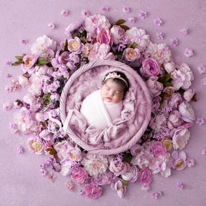 Könnte beinhalten: Ein neugeborenes Baby, eingewickelt in eine weiße Decke, mit einem kleinen Krönchen, liegt in einem geflochtenen Korb. Der Korb ist von einer Anordnung aus rosa und lila Blumen auf einem hellvioletten Hintergrund umgeben. Ein schönes florales Neugeborenen-Foto-Requisit.