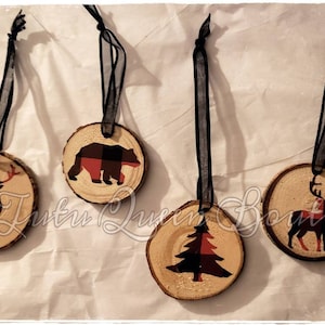 May include: Four rustic wood slice ornaments with black ribbon hangers. Each ornament features a different design: a deer, a bear, a Christmas tree, and another deer, all with red and black plaid accents. Ideal for festive decorations.