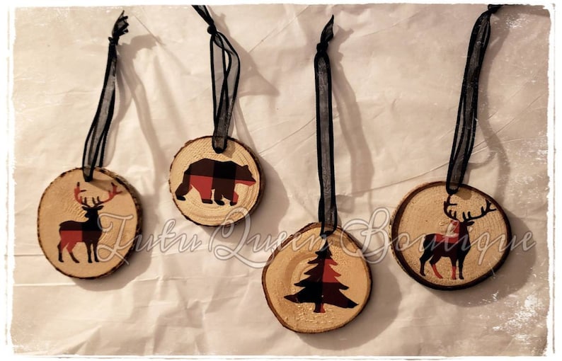 May include: Four rustic wood slice ornaments with black ribbon hangers. Each ornament features a different design: a deer, a bear, a Christmas tree, and another deer, all with red and black plaid accents. Ideal for festive decorations.