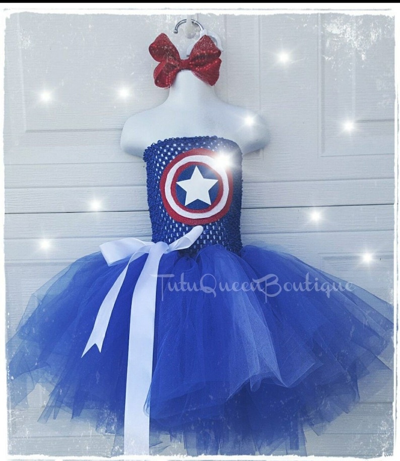 May include: Blue tulle tutu with a white satin ribbon and a red bow. The bodice is blue and white with a red, white, and blue Captain America shield.