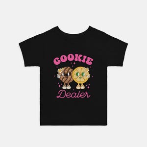 May include: Black t-shirt with a pink graphic of a cookie and a lemon slice holding hands. The text "Cookie Dealer" is written in pink below the graphic.
