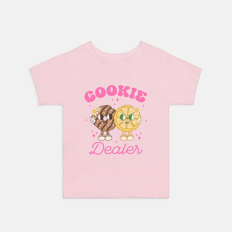 May include: Pink t-shirt with a cartoon cookie and lemon holding hands. The text "COOKIE DEALER" is printed in pink letters.