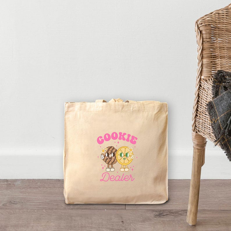 May include: A beige tote bag with a pink graphic of a cookie and a lemon slice with the text "COOKIE DEALER".