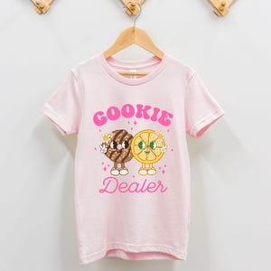 May include: Pink t-shirt with a graphic of a cookie and a lemon slice with the text "COOKIE DEALER" in pink.