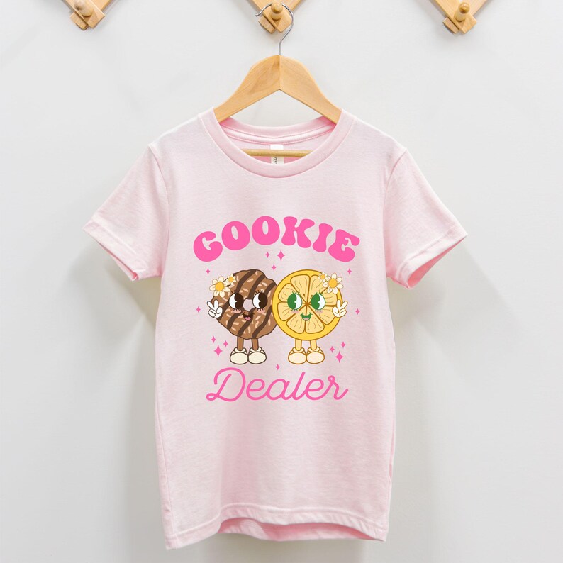 May include: Pink t-shirt with a graphic of a cookie and a lemon slice with the text "COOKIE DEALER" in pink.
