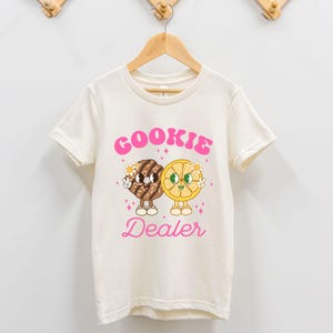 May include: A white t-shirt with a pink graphic of a cookie and a lemon slice with the text "COOKIE DEALER" in pink.