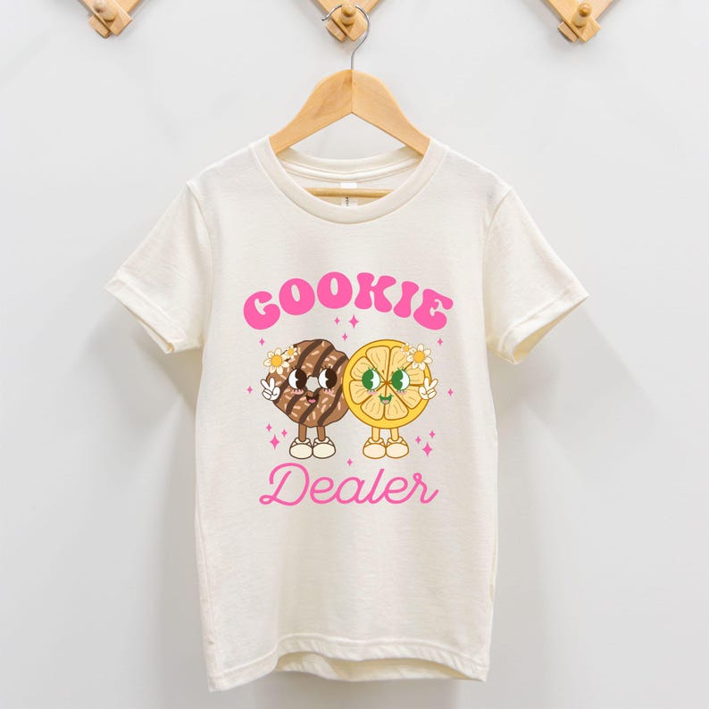May include: A white t-shirt with a pink graphic of a cookie and a lemon slice with the text "COOKIE DEALER" in pink.