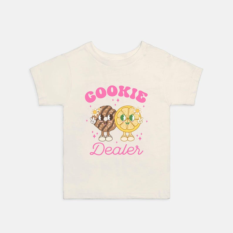 May include: A white t-shirt with a pink graphic of a cookie and a lemon slice holding hands. The text "Cookie Dealer" is written in pink below the graphic.