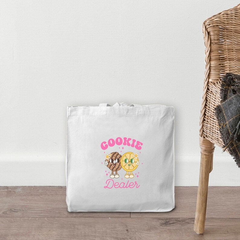 May include: White canvas tote bag with pink text that reads "COOKIE DEALER". The text is surrounded by two cartoon characters, a cookie and a lemon, both with smiling faces.