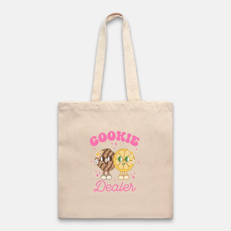 May include: A beige tote bag with a pink graphic of two cartoon cookies holding hands. The text "COOKIE DEALER" is written in pink letters.