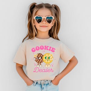 May include: A young girl wearing a light beige t-shirt with a pink graphic of a cookie and a lemon slice with the text "COOKIE DEALER". She is wearing heart-shaped sunglasses and has her hair pulled back in pigtails.
