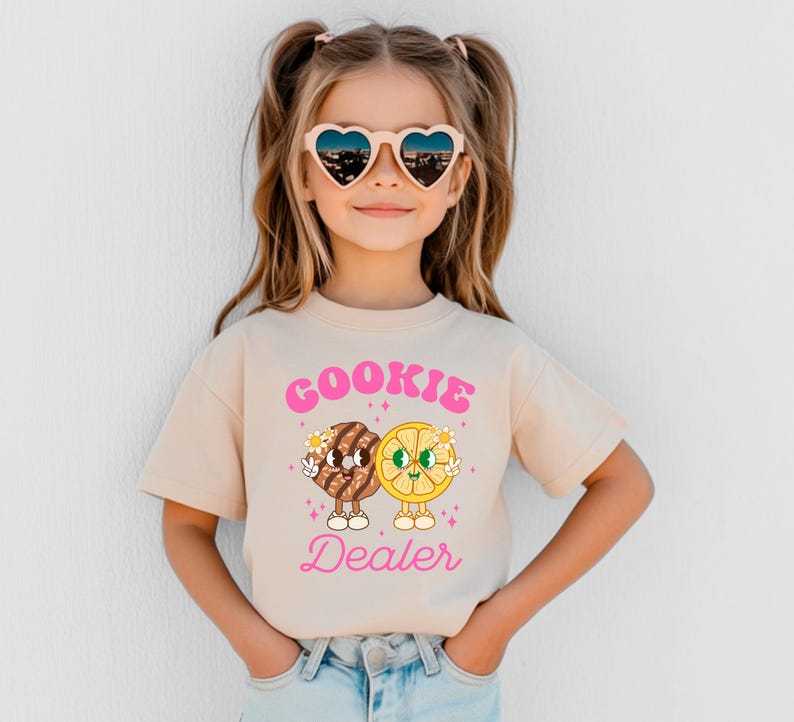 May include: A young girl wearing a light beige t-shirt with a pink graphic of a cookie and a lemon slice with the text "COOKIE DEALER". She is wearing heart-shaped sunglasses and has her hair pulled back in pigtails.