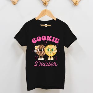 May include: Black t-shirt with a pink graphic of two cartoon cookies, one chocolate chip and one lemon, holding hands and the text "COOKIE DEALER".