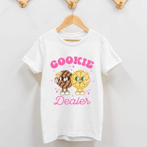 May include: White t-shirt with a pink graphic of a cookie and a lemon slice with the text "COOKIE DEALER" in pink.