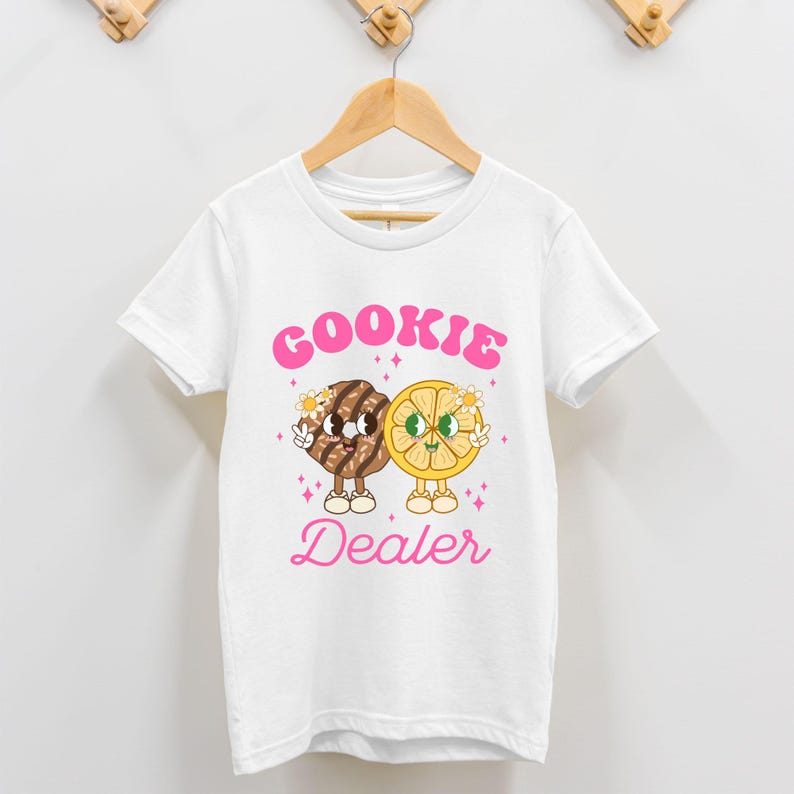 May include: White t-shirt with a pink graphic of a cookie and a lemon slice with the text "COOKIE DEALER" in pink.