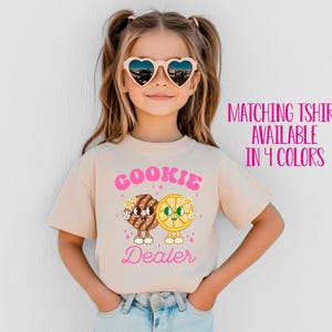 May include: A young girl wearing a light beige t-shirt with a pink graphic of a cookie and a lemon slice with the text "Cookie Dealer". She is wearing heart-shaped sunglasses and has her hair pulled back in pigtails.