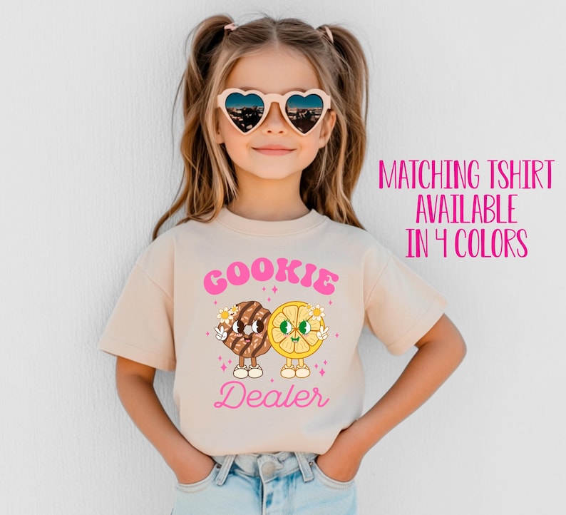 May include: A young girl wearing a light beige t-shirt with a pink graphic of a cookie and a lemon slice with the text "Cookie Dealer". She is wearing heart-shaped sunglasses and has her hair pulled back in pigtails.