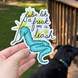 May include: A sticker featuring a turquoise worm-like creature with the text "feelin like a freak on a leash" in black script. The sticker has a white border and yellow starburst accents. The background includes a blurred view of a black dog and outdoor setting.