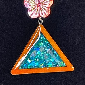 Puede incluir: Un collar de cordón de cuero marrón con un dije de flor rosa iridiscente y un dije triangular lleno de purpurina turquesa.