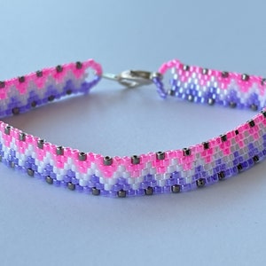 May include: A beaded bracelet with a chevron pattern in shades of pink, purple, and white with silver accents.