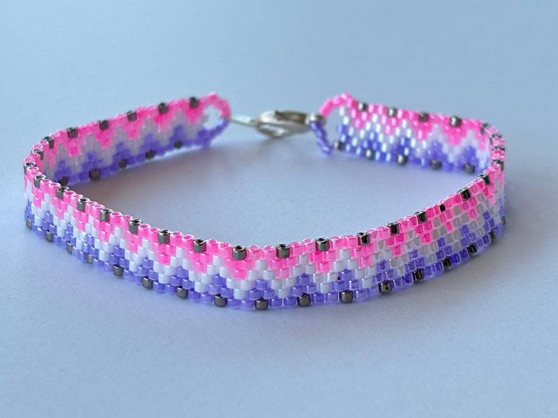 May include: A beaded bracelet with a chevron pattern in shades of pink, purple, and white with silver accents.